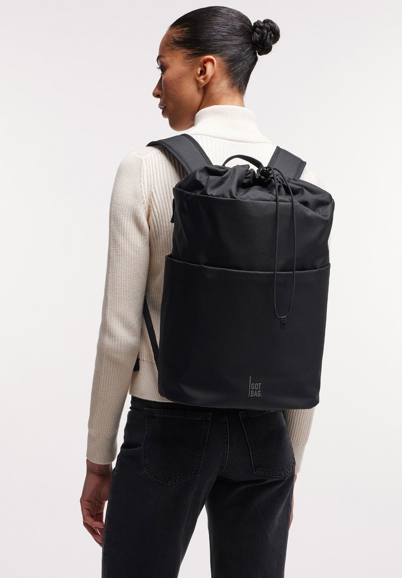 Nainen nutturalla, pukeutuneena vaaleaan pooloneuleeseen ja mustiin farkkuihin, kantaa suurta mustaa narulla suljettavaa reppua, jossa on "GOT BAG"-logo.