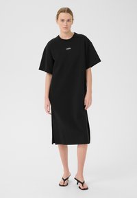 Czarna, oversize sukienka t-shirtowa z krótkimi rękawami, okrągłym dekoltem, bocznymi rozcięciami i subtelnym detalem z logo. Gładka struktura materiału.