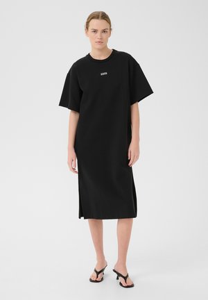Zwart oversized t-shirt jurk met korte mouwen, ronde hals, zijslitsen en subtiele logodetail. Soepele stoftextuur.