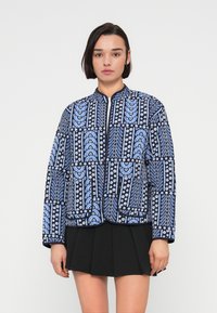 Μπλε καπιτονέ μπουφάν με σχέδια λουλουδιών και γεωμετρικά μοτίβα, με ψηλό γιακά και φερμουάρ μπροστά. Το υλικό φαίνεται μαλακό και υφασμένο.