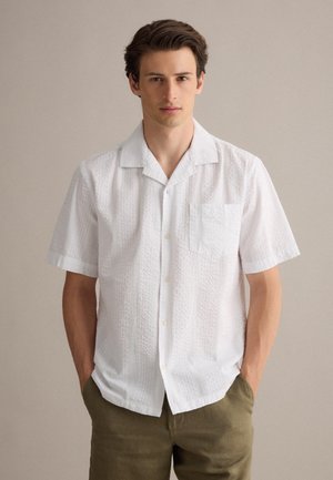 Junger Mann mit dunklen Haaren, der ein weißes, strukturiertes Kurzarm-Hemd mit Knöpfen und olivgrünen Hosen trägt, steht mit den Händen in den Taschen.