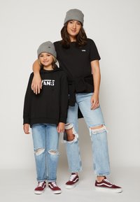 Due persone indossano berrette di maglia grigia abbinate. L'adulto ha una maglietta nera e jeans chiari strappati, mentre il bambino indossa una felpa nera e jeans chiari. Entrambi indossano sneaker rosse e bianche.