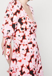 Kukallinen mekko, jossa on vaaleanpunainen tausta punaisilla ja mustilla kukkakuviolla, syvä V-nukka, pullottavat hihat ja nauhakoristeet hihoissa.