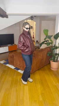 Bordo odos bombardieriaus striukė su užtrauktuku, tamsios džinsai ir šviesios spalvos batai. Vidaus aplinka su augalu ir sofa fone.