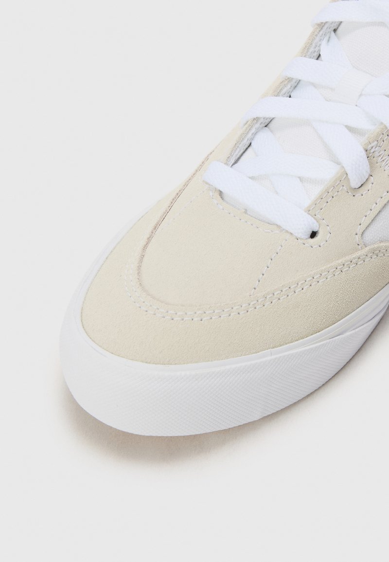 Zapatilla de ante beige con cordones blancos y suela de goma. Presenta un detalle de costura y una textura suave en la parte superior.