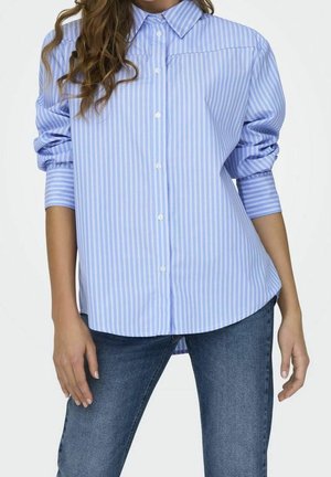 Femme portant une chemise à rayures verticales bleu clair à boutons avec les manches retroussées et un jean bleu, debout devant un fond uni.