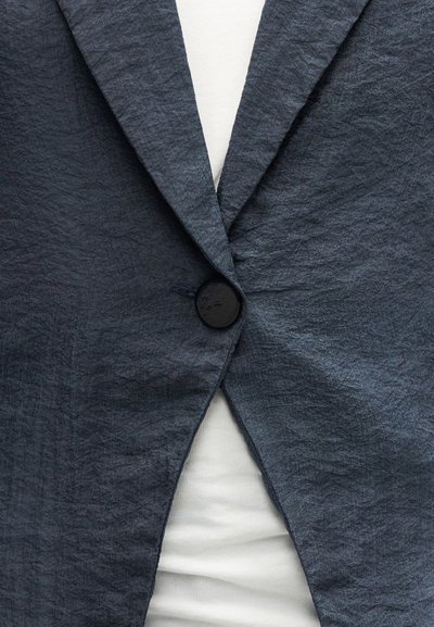 Približni prikaz tamnosivog naboranog blejzera zakopčanog preko bijele košulje, ističući teksturu tkanine i detalje na dugmetu.