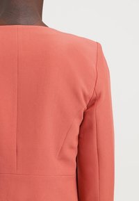 Blazer sur mesure couleur rouille avec une texture lisse, des épaules structurées et des lignes épurées, au design simple et minimaliste.