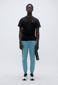 Μαύρο κοντομάνικο μπλουζάκι, τιρκουάζ αθλητικές παντελόνες και μαύρα αθλητικά παπούτσια. Το μοντέλο κρατάει ένα μαύρο μπουκάλι νερού. Απλός και αθλητικός σχεδιασμός.