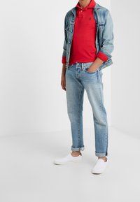 Denim takki, punainen polo paita, vaaleansiniset farkut, valkoiset slip-on kengät. Rentoa asua, jossa farkkujen hihat on käännetty ja hienovarainen logo-yksitys.