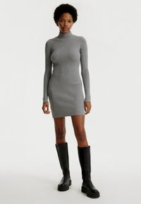 Graues geripptes Strickkleid mit hohem Kragen, langen Ärmeln und einer figurschmeichelnden Silhouette. Kombiniert mit schwarzen knielangen Stiefeln und minimalistischer Gestaltung.