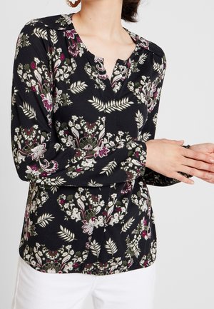 Kvinde iført sort bluse med lange ærmer og blomsterprint i hvid, grøn og lilla, kombineret med hvide bukser, hænderne foldet foran.