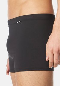 Schwarze Badehosen aus dehnbarem Material, mit einem schlichten Design, niedrigem Bund und einem Etikett auf der Rückseite. Glatte Textur.