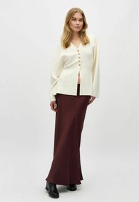 Cardigan di maglia crema con scollo a V e bottoni in legno, abbinato a una lunga gonna bordeaux. L'outfit è completato da stivaletti neri alla caviglia.