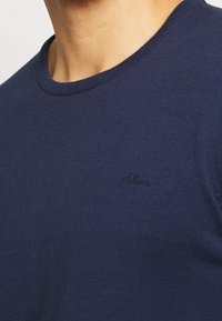 Mörkblå rundhalsad tröja i ribbad tyg, med en diskret präglad logotyp på bröstet som visar en slät, strukturerad yta.