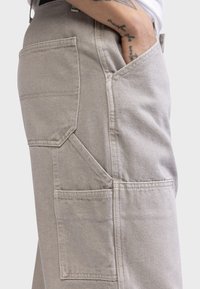 Beige cargo pants with multiple pockets, visible stitching, and a sturdy fabric texture. Features a high waist and tapered leg design.