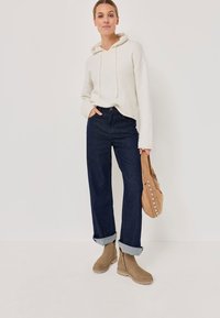 Femme portant un hoodie crème, un jean foncé retroussé, des bottines beiges, tenant un sac à main à studs beiges devant un fond clair et uni.