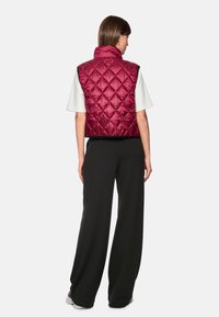 Burgundy quilted sleeveless vest with a high collar, paired with a white t-shirt and black wide-leg pants, shown from the back.