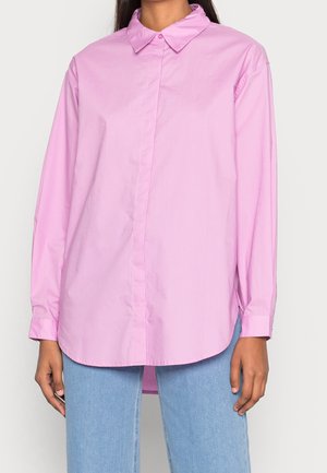 Mujer con camisa rosa de manga larga y botones, de corte suelto y con cuello, combinada con jeans azul claro, sobre un fondo liso.