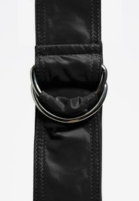 Czarny materiałowy pasek z okrągłym srebrnym pierścieniem. Gładka tekstura z widocznymi detalami przeszycia wzdłuż krawędzi.