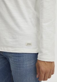 Hvid langærmet skjorte i et let, struktureret stof, med et lille mærke på kanten med tekst, parret med blå denim jeans.