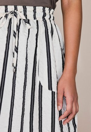 Nærbillede af en hånd, der hviler ved siden af en hvid nederdel med lodrette sorte striber og elastisk linning med bindebånd.