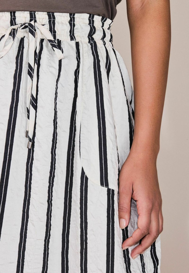 Nahaufnahme einer Hand, die neben einem weißen Rock mit vertikalen schwarzen Streifen und einem gebundenen elastischen Bund ruht.