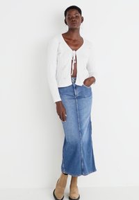 White quilted cardigan with tie front, paired with a long blue denim skirt. Exhibits a fitted design and light texture. Brown heeled boots complete the look.