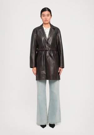 Femme portant un manteau en cuir marron foncé ceinturé, une chemise blanche à motifs, un jean évasé bleu clair et des chaussures noires à bout pointu, se tenant devant un fond uni.