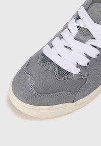 Sneaker in suede grigio con lacci bianchi, dettagli in rete testurizzata e suola in gomma crema, con cuciture arcuate lungo la parte superiore.