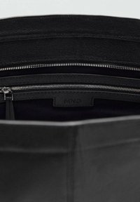 Intérieur d'un sac en cuir noir avec une texture lisse, plusieurs compartiments, une poche zippée et une étiquette de marque. Doublure en tissu sombre.