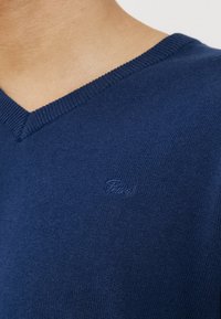 Close-up of a person wearing a navy blue V-neck sweater with a small, subtle embroidered logo on the chest.