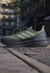 Olivgrüner und schwarzer Adidas-Laufschuh mit dicker Sohle, drei grünen Streifen an der Seite, auf Steinpflaster in städtischer Außenumgebung platziert.