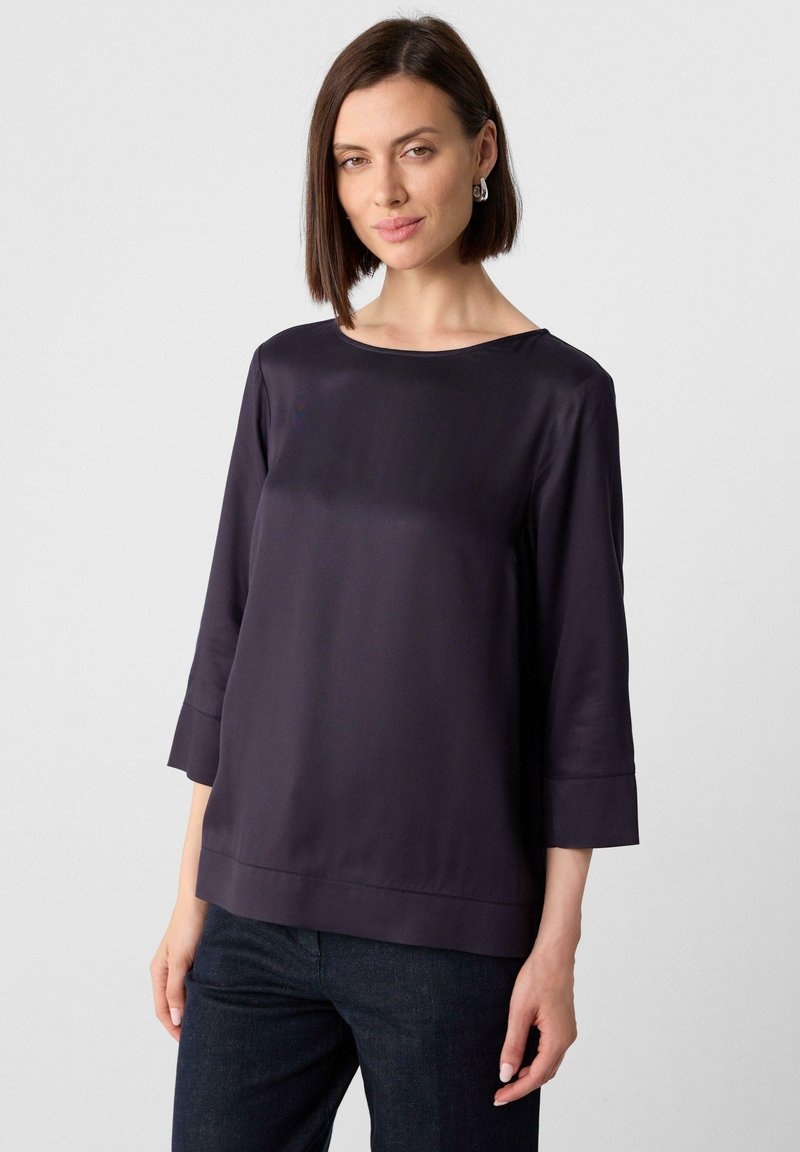 Top in satin viola scuro con scollo a barca, maniche a tre quarti e orlo dritto. Abbinato a pantaloni in denim scuro, con una texture liscia.