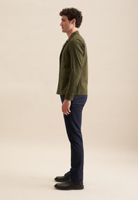 Olivgrüne Jacke mit strukturiertem Design, einreihig, zwei Vordertaschen, kombiniert mit navyblauen Hosen und schwarzen Schuhen. Einfache Texturen.