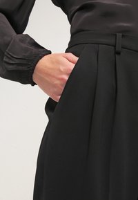 Svart skräddarsydda byxor med veck, med en sidoficka och en slät yta. En långärmad svart blus syns ovanför.