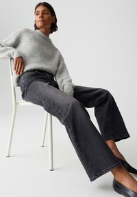Pull gris côtelé associé à un jean large en denim foncé. Le jean a une taille haute, cinq poches et une légère décoloration. Modèle assis sur une chaise.