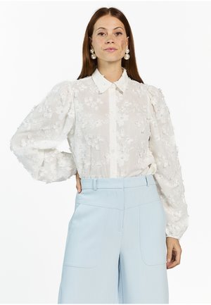 Frau mit langem braunem Haar trägt eine weiße Bluse mit Blumenstickerei, Puffärmeln und hellblauen, hoch taillierten Hosen mit weitem Bein.