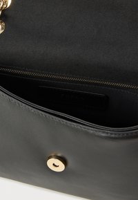 Čierna kožená taška s magnetickým zapínaním, kovaním v zlatej farbe, hladkou textúrou a vnútornou etiketu s označením značky. Vo vnútri je vrecko na zips.