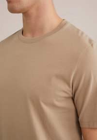 Camiseta de algodón beige con cuello redondo y mangas cortas, que presenta una textura suave y un diseño discreto. Sin logotipos ni patrones visibles.
