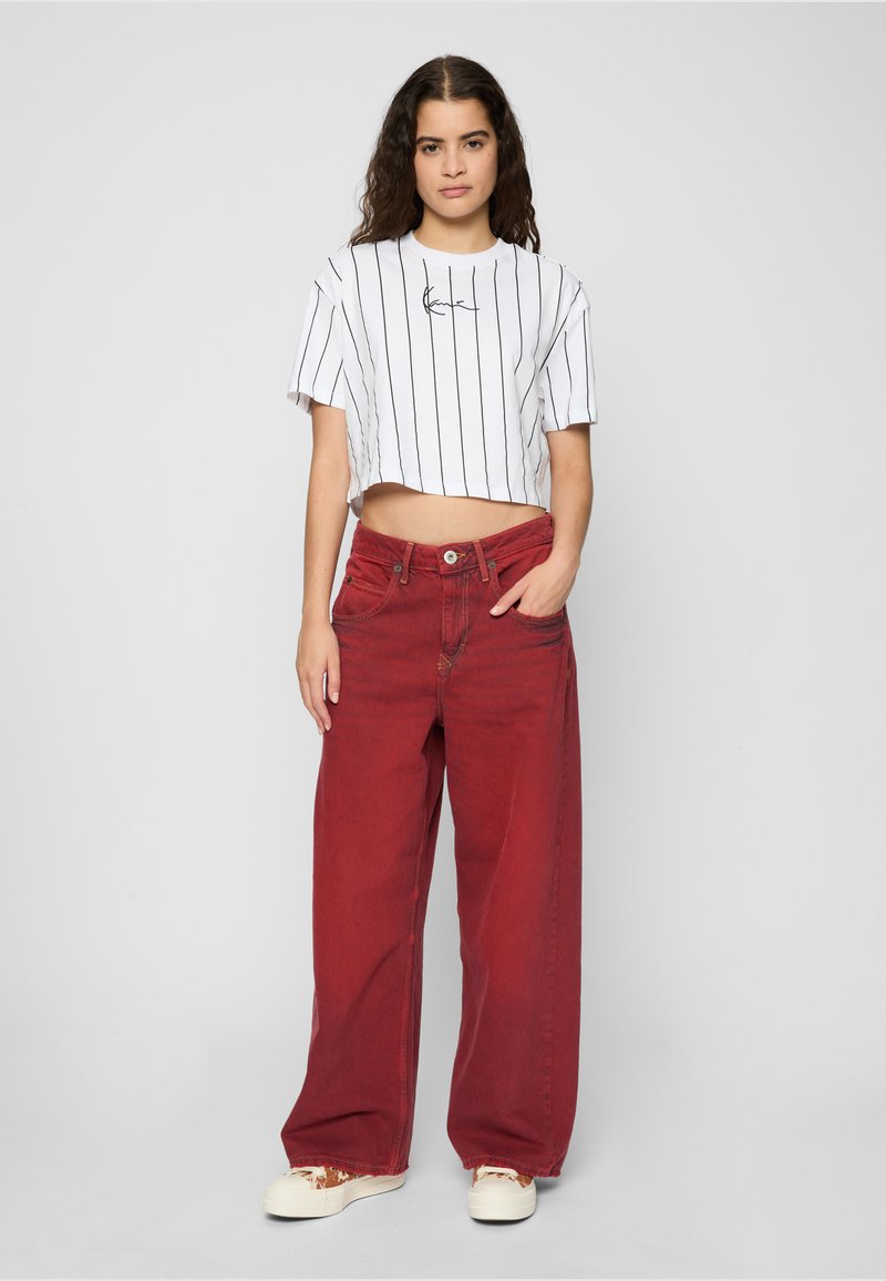 Tânără cu păr lung purtând un tricou alb scurt cu dungi, pantaloni largi roșii și pantofi sport albi, stând cu o mână în buzunar.