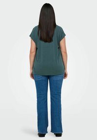 Green, short-sleeved top with a loose fit, paired with flared blue jeans and black shoes, viewed from the back. Smooth fabric texture.