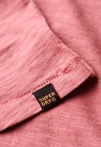 Tessuto rosa testurizzato con cuciture visibili, con un'etichetta nera che riporta la scritta "SUPER DRY" in lettere dorate sul bordo.