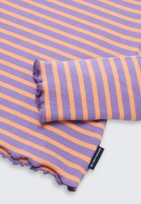 Fioletowo-pomarańczowa bluzka z długim rękawem w paski o prążkowanej fakturze, z falbaną u dołu i metką z logo w prawym dolnym rogu.