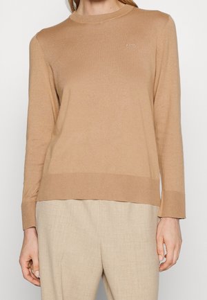 Femme portant un pull à manches longues couleur camel avec poignets et ourlet côtelés, associé à un pantalon beige clair, debout devant un fond uni.