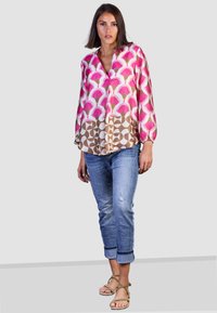 Bunte Bluse mit pinken und braunen geometrischen Mustern, langen Ärmeln, V-Ausschnitt, kombiniert mit hellblauen, ausgewaschenen Jeans und beige Sandalen.