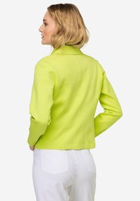 Limoengroene getailleerde jas met een gestructureerde kraag, lange mouwen en naaddetails, gecombineerd met witte broek. Een gladde, lichte stof.