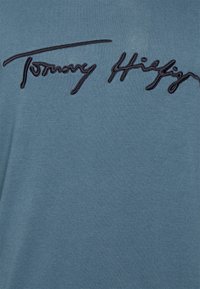 T-shirt in cotone blu con un'importante firma ricamata nera di "Tommy Hilfiger" sul petto. Texture morbida, design casual.