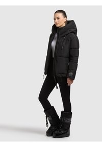 Chaqueta acolchada negra con cuello alto, bolsillos con cremallera y dobladillo con cordón. Combinada con leggings negros y botas de invierno negras de gran tamaño.