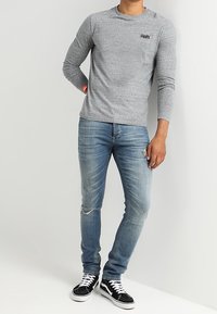 Harmaa pitkähihainen t-paita pyöreällä pääntiellä ja kirjailulla logolla, yhdistetty vaaleansinisillä, kapealahkeisilla kulutetuilla farkuilla ja mustilla tennareilla.